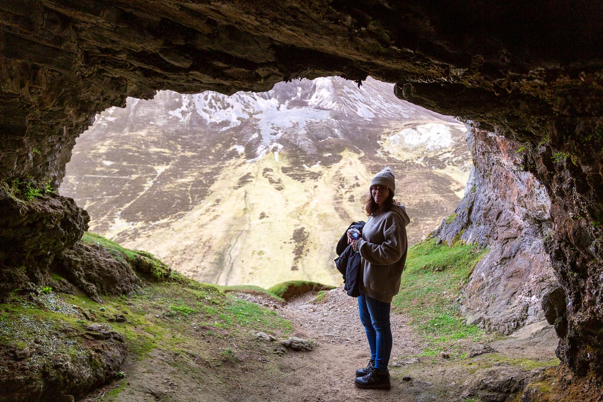 Janette standing inside the entrance of one of the Bone Caves at Inchnadamph with limestone walls framing a view of the Assynt mountains beyond