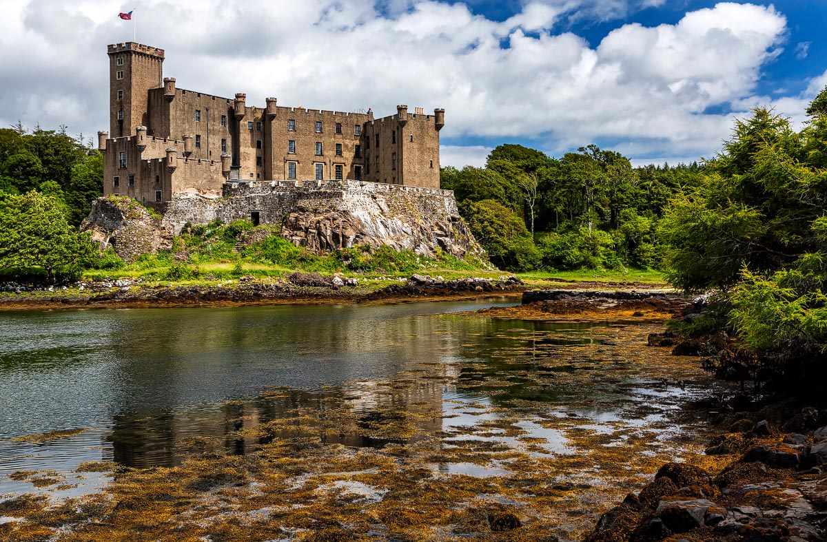 Dunvegan Castle reflected in water with seaweed-fringed shores in the foreground and trees behind